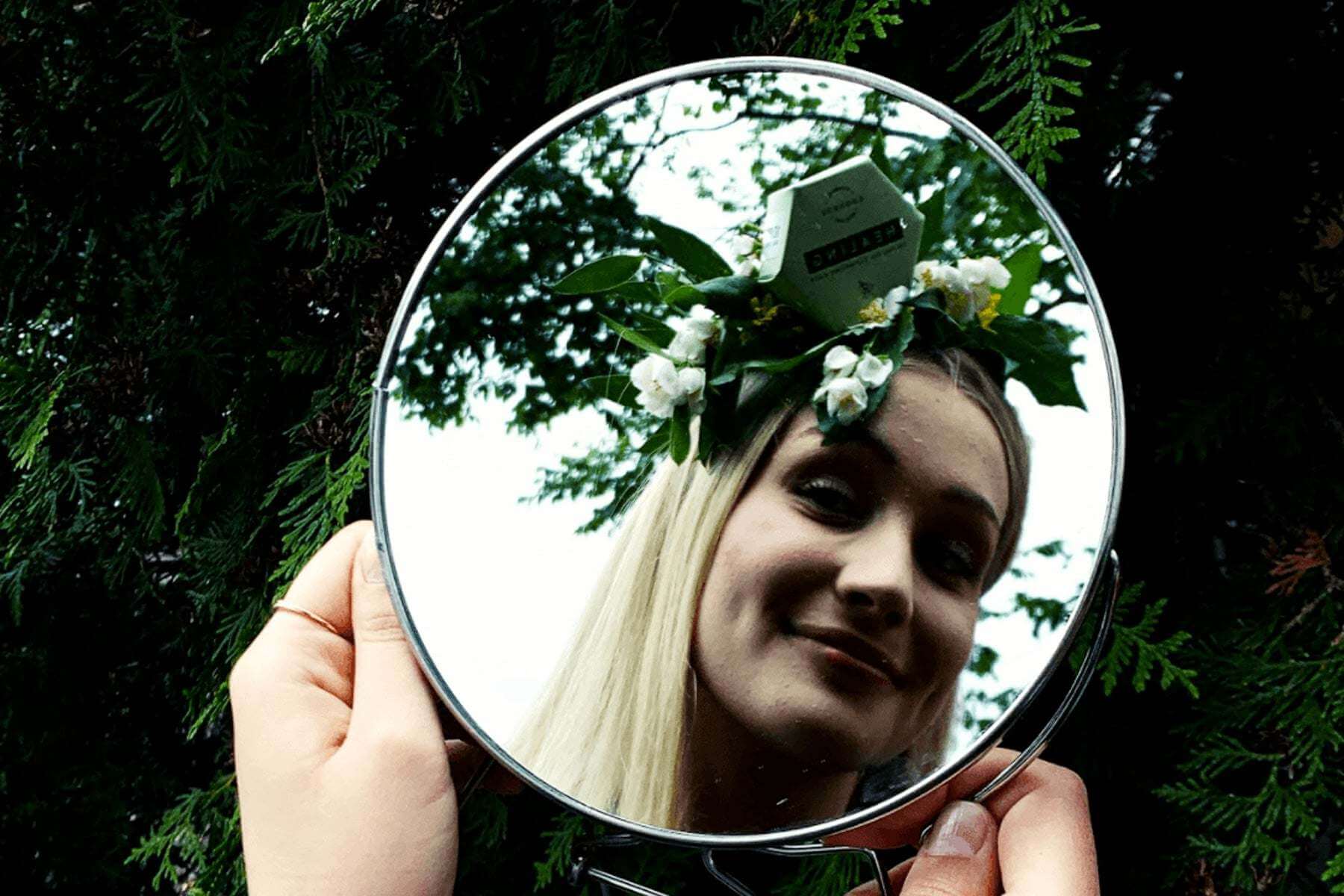 teen with clear skin holding a round mirror outdoors reflecting her face with a floral headband, representing gentle skincare for teenage skin