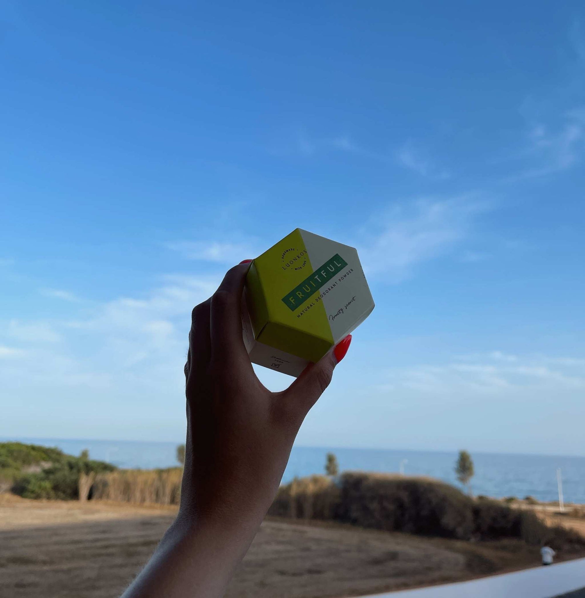 Hand holding a small natural skincare product box against a clear blue summer sky and coastal landscape, highlighting kesäihonhoito summer skincare.