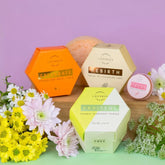 Kesän hehku ihonhoitosetti with natural skincare products including oil cleansing cake, sparkling body cake, natural deodorant powder, tinted lip balm, and eco-friendly facial sponge surrounded by flowers.
