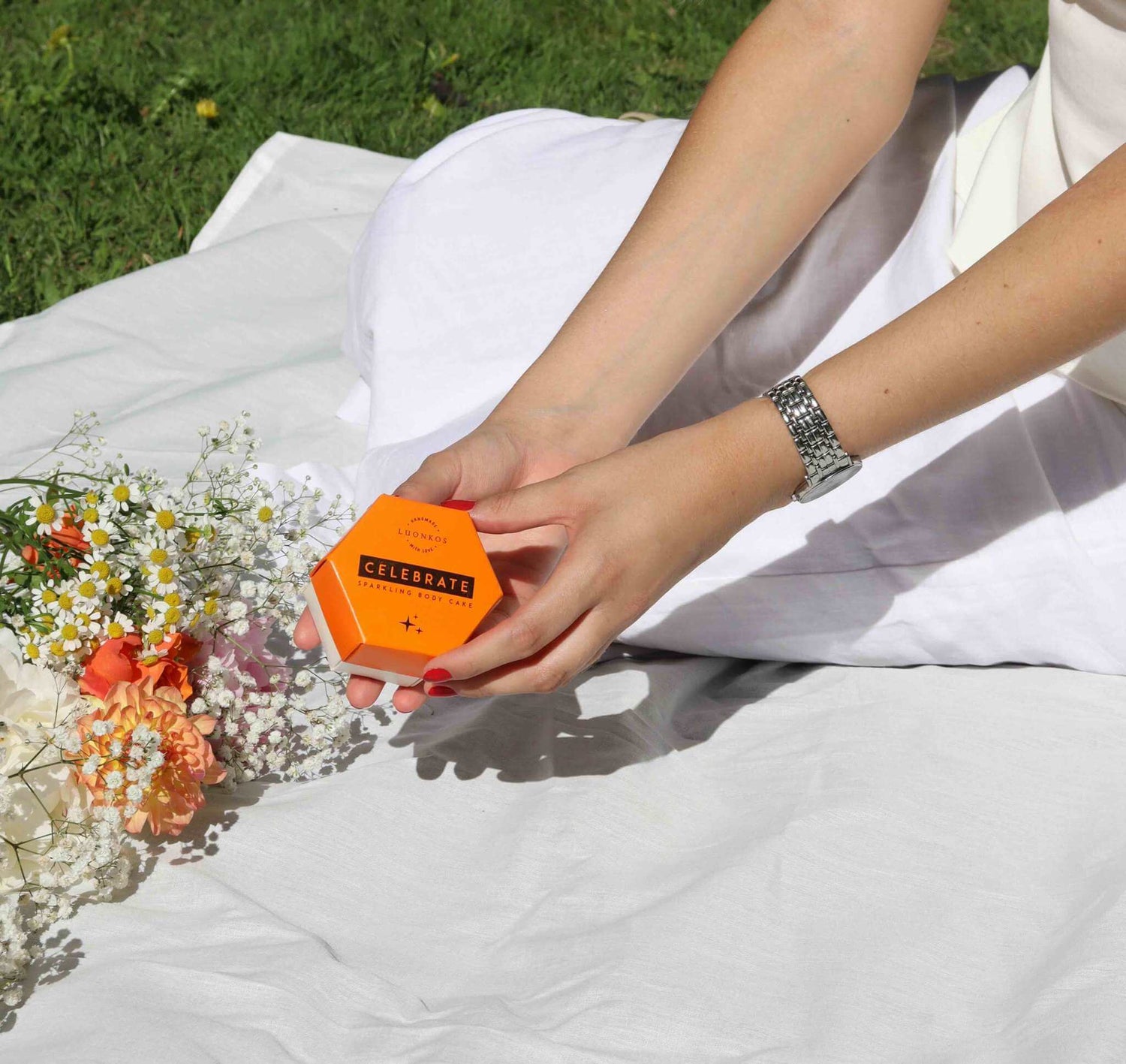 Hand holding orange Luonkos Celebrate multi-use beauty product box on white cloth with flowers nearby in outdoor setting