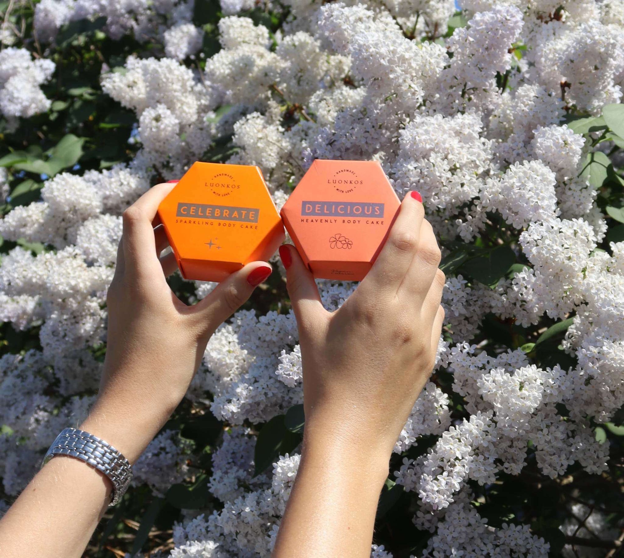 Hands holding Luonkosin Hehku sarja Celebrate and Delicious hexagonal body oil cakes in front of white flowering bush