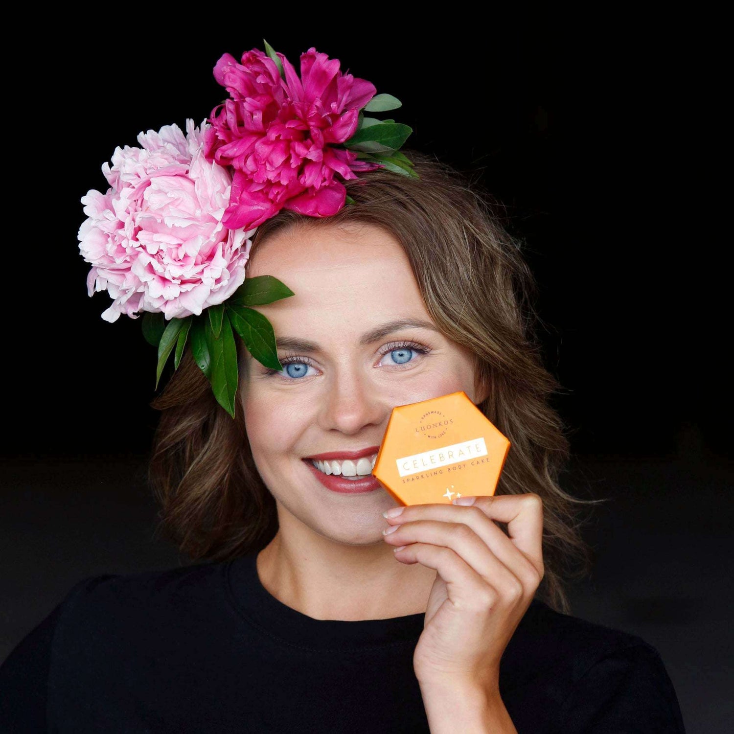 Smiling woman with flowers in hair holding natural skincare product Celebrate by Luonkos
