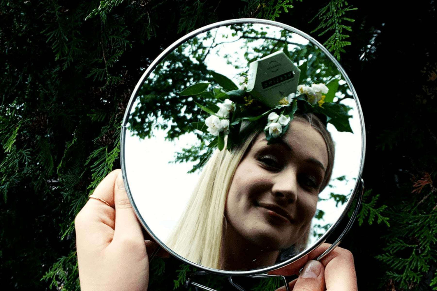 teen with clear skin holding a round mirror outdoors reflecting her face with a floral headband, representing gentle skincare for teenage skin