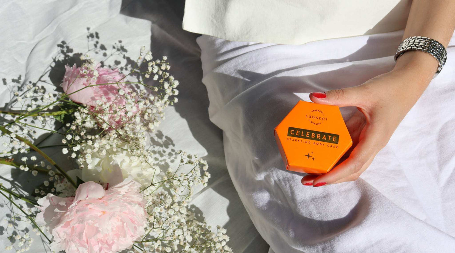 Hand holding Luonkos Celebrate sparkling body cake in orange hexagonal packaging on white fabric next to pink and white flowers, summer beach essentials.