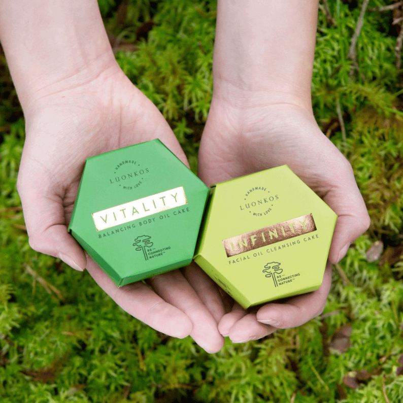 Hands holding Luonkos Vitality balancing body oil cake and Infinity facial oil cleansing cake on green moss background, Luonkos tuotteet allergiatunnus safe skincare products for sensitive skin.