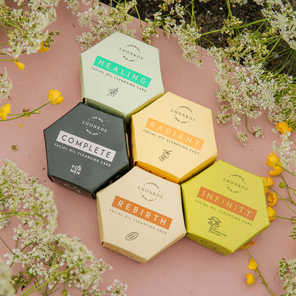 Five hexagonal boxes of Luonkos facial oil cleansing cakes in various colors labeled Healing, Radiant, Complete, Rebirth, and Infinity surrounded by delicate flowers on a pink surface representing natural skincare and oil cleansing.